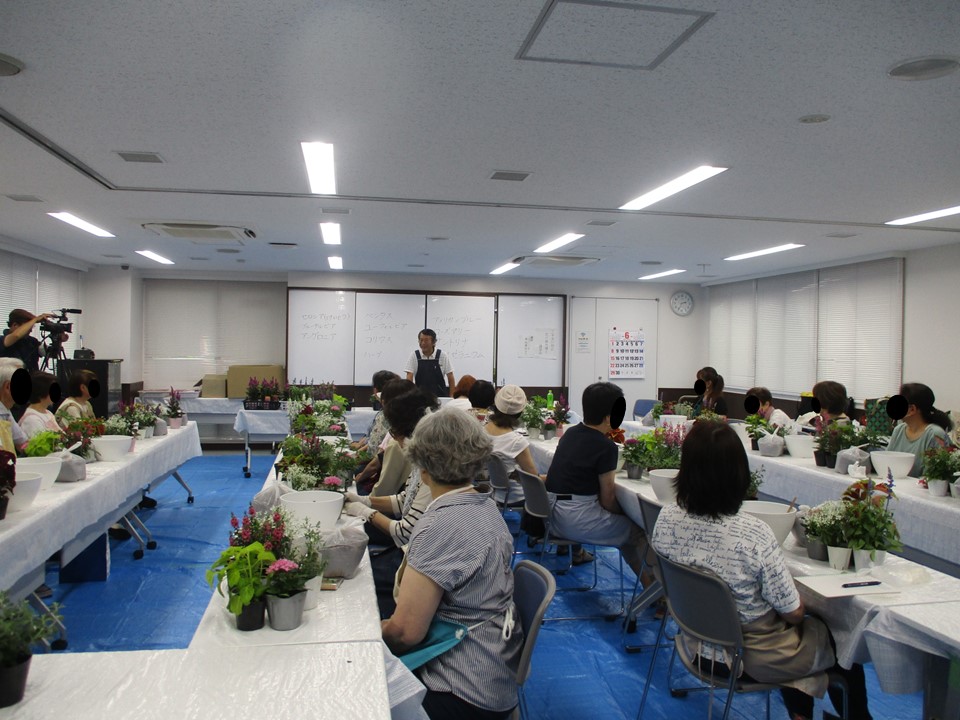 講習会の様子、物品一式が用意しているテーブルの前に参加者が座り、前方で説明する井出さんの話を聞いている